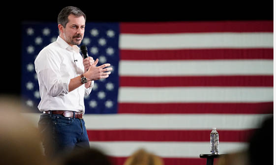 Pete Buttigieg standing in front of a large American flag.
